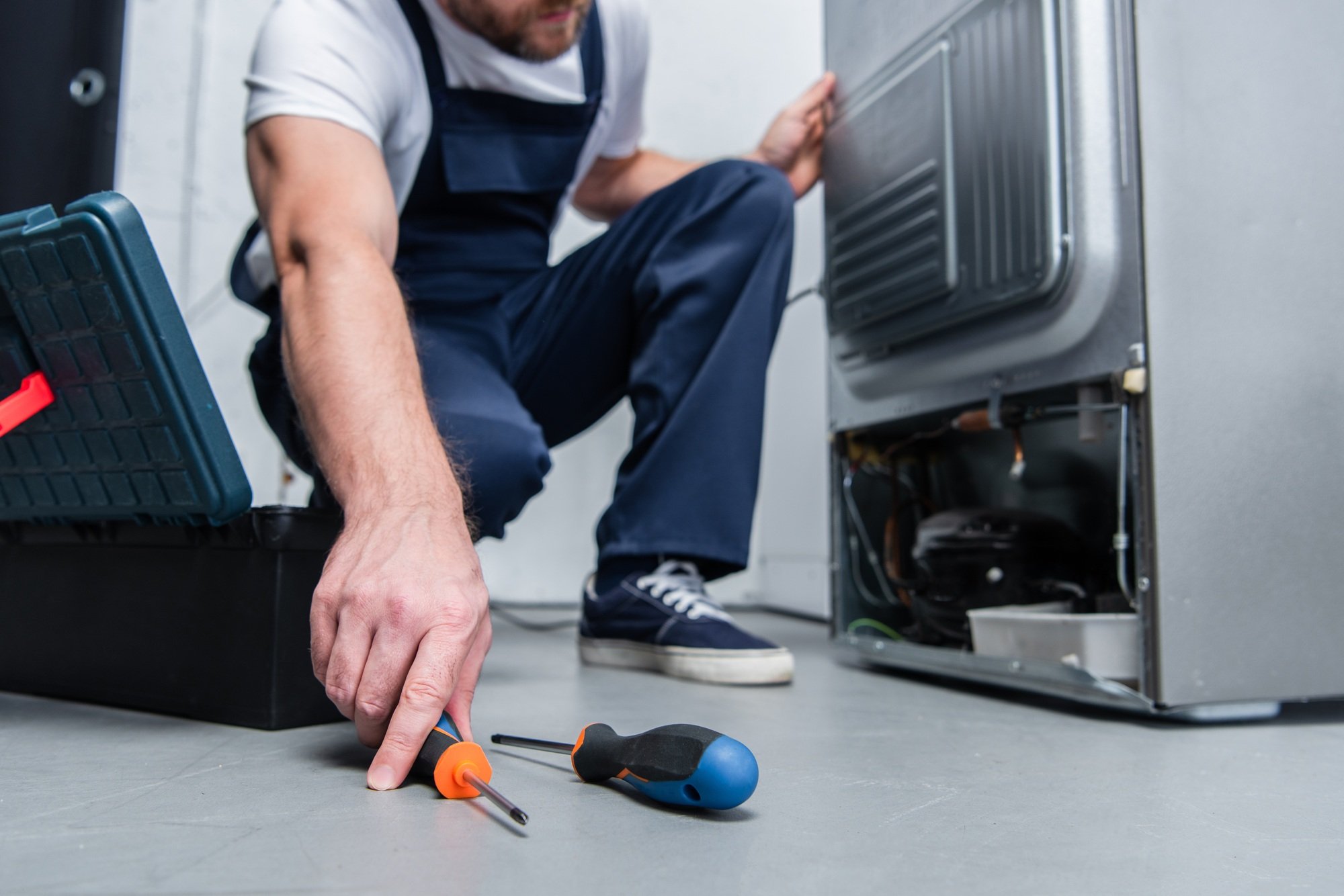 cropped shot of repairman in working overall taking screwdriver from floor near broken refrigerator
